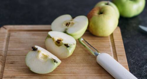 Geviertelte Äpfel mit Schäler auf Schneidebrett mit dunkelgrauem Hintergrund, von oben seitlich fotografiert.
