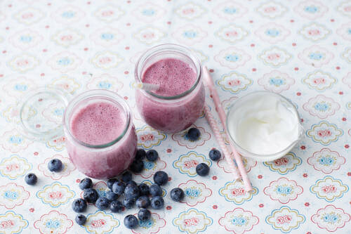 Apfel-Heidelbeer Smoothies in Gläsern mit Strohhalmen neben Heidelbeeren und Joghurt auf bunter Tischdecke.