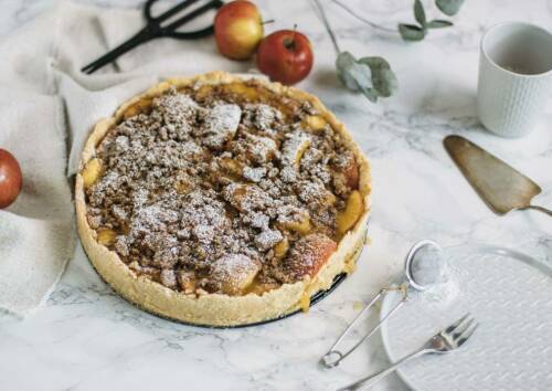 Apfel-Streuselkuchen auf weißem Hintergrund, daneben Äpfel und Tortenheber, Puderzucker. Von oben fotografiert.