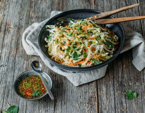 Asiatischer Chinakohlsalat in schwarzem Teller mit Holzstäbchen neben würzigem Dressing auf Holztisch.