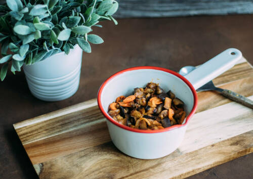 Caponata aus Auberginen in einem kleinen Topf auf einem Holzbrett, von oben fotografiert.