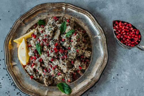 Auberginensalat mit Tahin auf Metallteller neben Granatapfelkernen vor hellem Hintergrund,