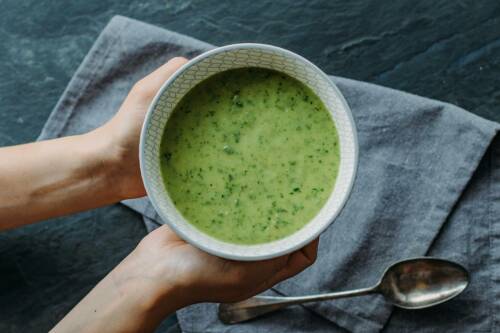 Avocado-Minze-Suppe in Schüssel mit Händen vor dunklem Hintergrund.