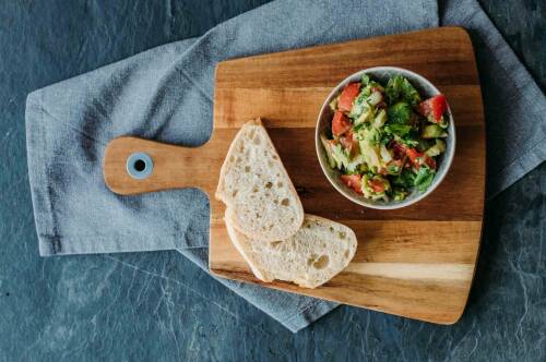 Avocado-Salsa in Schüssel auf Schneidebrett neben aufgeschnittenem Brot vor dunklem Hintergrund.