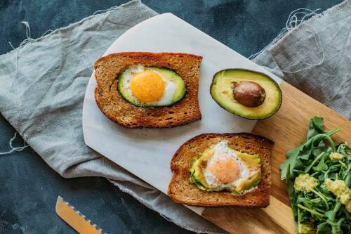 Avocado-Spiegelei-Toast neben Avocado und Rucola auf Schneidebrett vor dunklem Hintergrund.