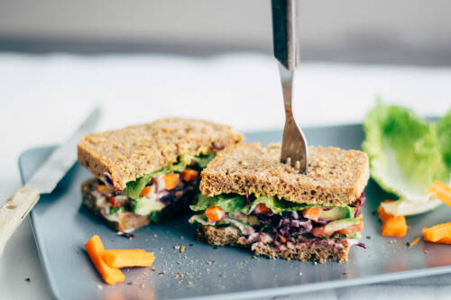 Avocado Veggie Deluxe Sandwich auf Teller mit Gabel und Salat vor hellem Hintergrund.