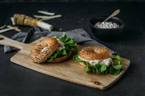 Bagel mit Apfel-Meerrettich-Aufstrich und Feldsalat auf Schneidebrett vor dunklem Hintergrund.