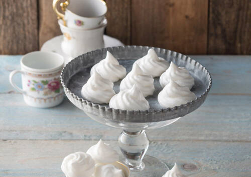 Baiser-Tupfen auf einer Servierplatte, Teetassen im  Hintergrund, von vorne fotografiert.