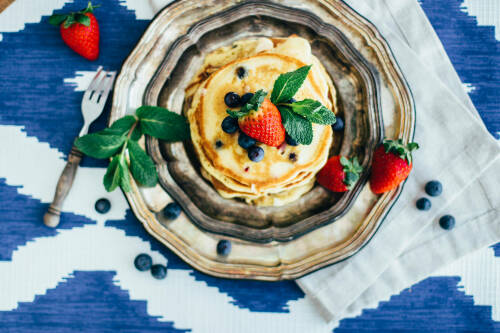 Beerige Pancakes auf Metallteller neben Beeren vor hellem Hintergrund.