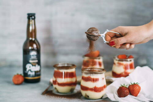 Bieramisu mit Erdbeersoße in Schälchen mit Hand neben Bier und Erdbeeren vor hellem Hintergrund.