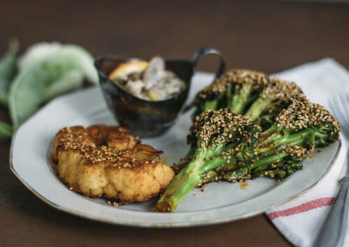 Mit Sesam panierte Blumenkohl- und Brokkolisteaks auf einem Teller, daneben ein Dip. Von der Seite fotografiert.