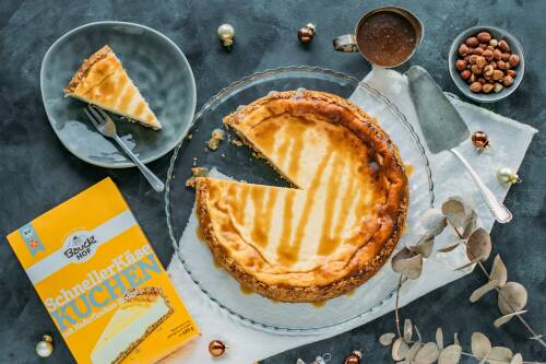 Frisch gebackener Bratapfel-Käsekuchen auf Glasplatte, ein Stück auf Teller daneben, daneben auch: Packung "Schneller Käsekuchen" von Bauckhof
