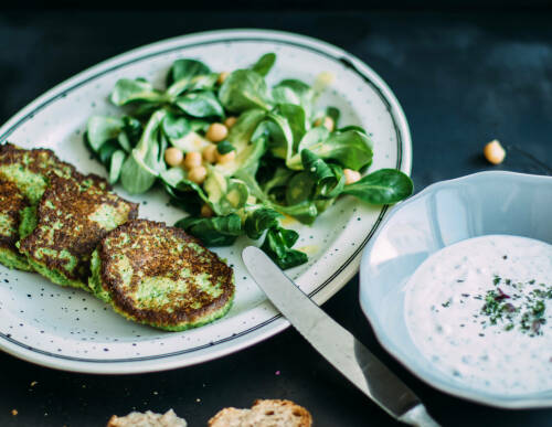 Brokkoli-Parmesan-Bratlinge mit Feldsalat und Dip auf einem weißen Teller auf dunklem Hintergrund, von oben fotografiert.