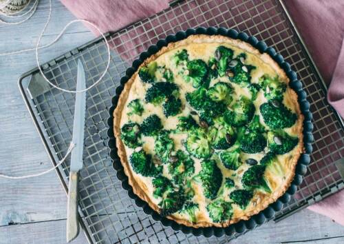 Leckere Brokkoliquiche frisch aus dem Ofen, noch in der Form auf einem Gitter, daneben ein Messer. Von oben fotografiert.