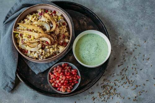 Bulgurpfanne mit Fenchel und Granatapfel in Schüsseln auf Serviertablett vor dunklem Hintergrund.
