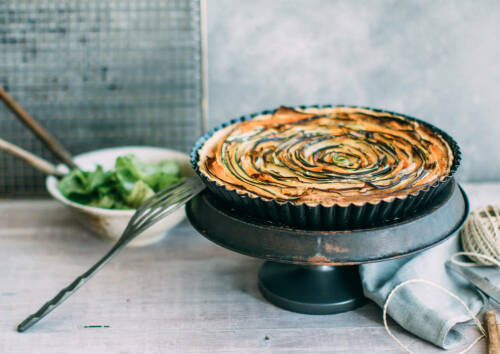 Gemüsetart im Spiral-Look mit Zucchini, Karotten und Auberginen dazu eine Schüssel mit grünem Salat im Hintergrund, von vorne fotografiert.