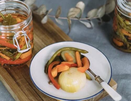 Buntes eingelegtes Gemüse in Weckglas und auf weißem Teller mit Löffel auf Schneidebrett vor hellem Hintergrund.