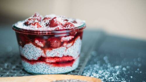 Kleine Weckglas gefüllt mit Chiapudding und Kirschkompott vor einem dunkelgrauen Hintergrund, darüber Puderzucker. Von der Seite fotografiert.