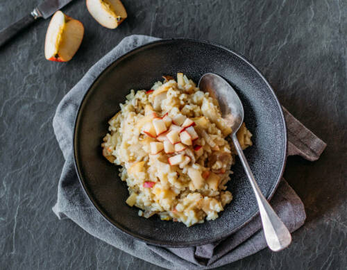 Chicoree-Risotto in schwarzem Teller mit Löffel neben aufgeschnittenem Apfel vor dunklem Hintergrund.