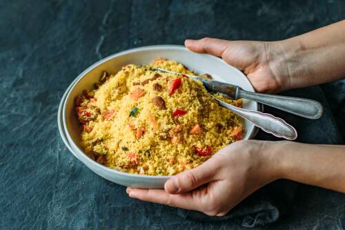 Helle, flache Schale gefüllt mit Couscous Salat, darin liegen noch Messer und Gabel. Dunkelgrauer Hintergrund, von oben fotografiert.