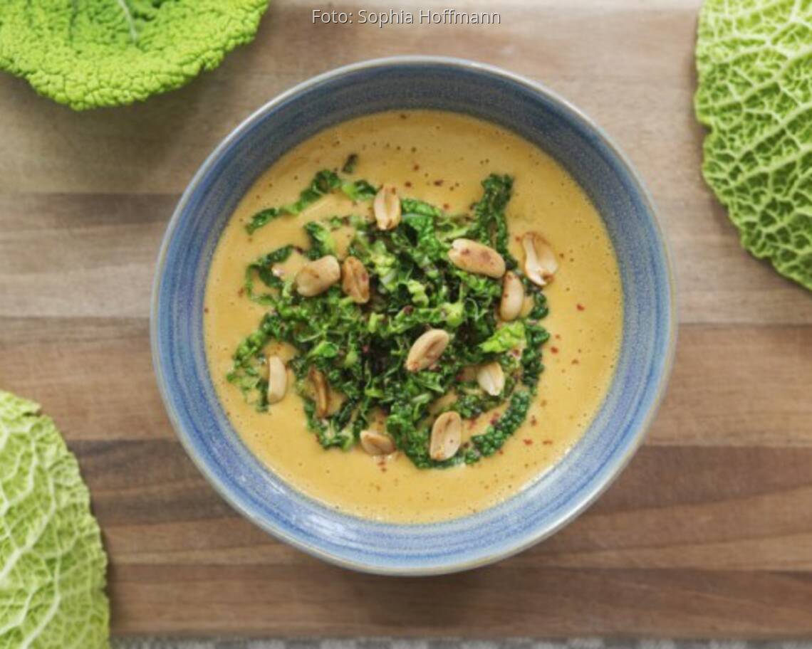 Wirsing-Erdnuss-Suppe in grauer Schale auf Holzbrett mit Wirsingblättern dekoriert