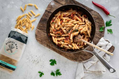Eine offene Packung Penne von DINKEL MAX neben einer Pfanne mit Penne all'Arrabbiata.