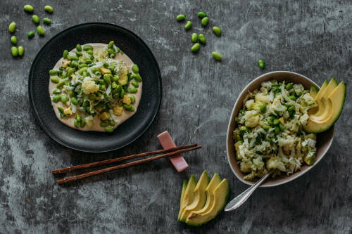 Edamame Avocado Salat in zwei Schüsseln neben Stäbchen und Edamame sowie Avocado vor dunklem Hintergrund.