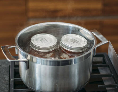 Zwei Weckgläser in kochendem Wasser eines großen Topfes auf Gasherd vor dunklem Hintergrund.