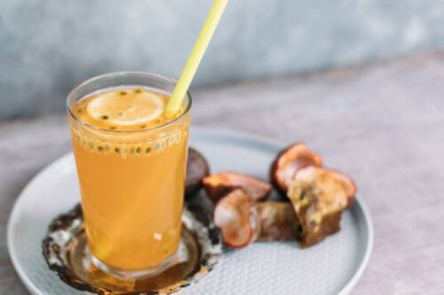 Glas Eistee mit Zitrone und Strohalm auf einem Teller mit Maracuja-Stücken vor hellem Hintergrund von vorne fotografiert