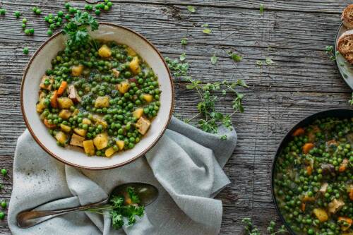Erbseneintopf mit Kräutern in weißem Teller neben Erbsen und Löffel vor hellem Hintergrund.