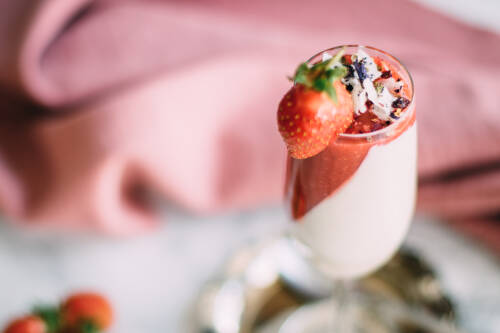 Sektglas gefüllt mit Cashew Panna Cotta und Erdbeer Mousse vor einem rosa Hintergrund von oben-seitlich fotografiert.