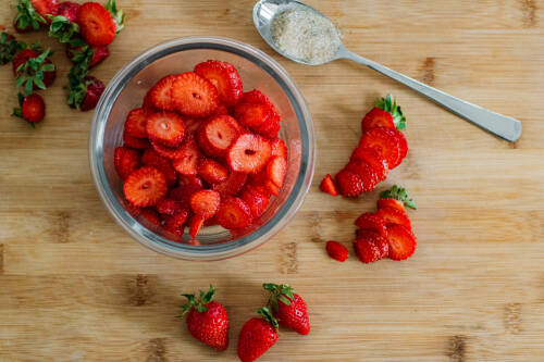 Erdbeeren geschnitten in Schüssel neben Löffel auf hellem Schneidebrett.