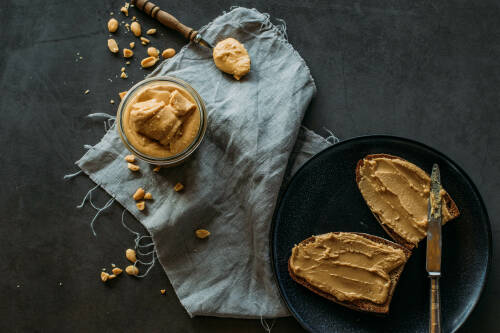 Zwei Erdnussbutterbrote auf schwarzem Teller neben Erdnussbutterglas und Löffel vor dunklem Hintergrund.