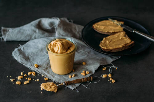 Erdnussbutter in Glas neben Löffel und zwei geschmierten Broten vor dunklem Hintergrund.