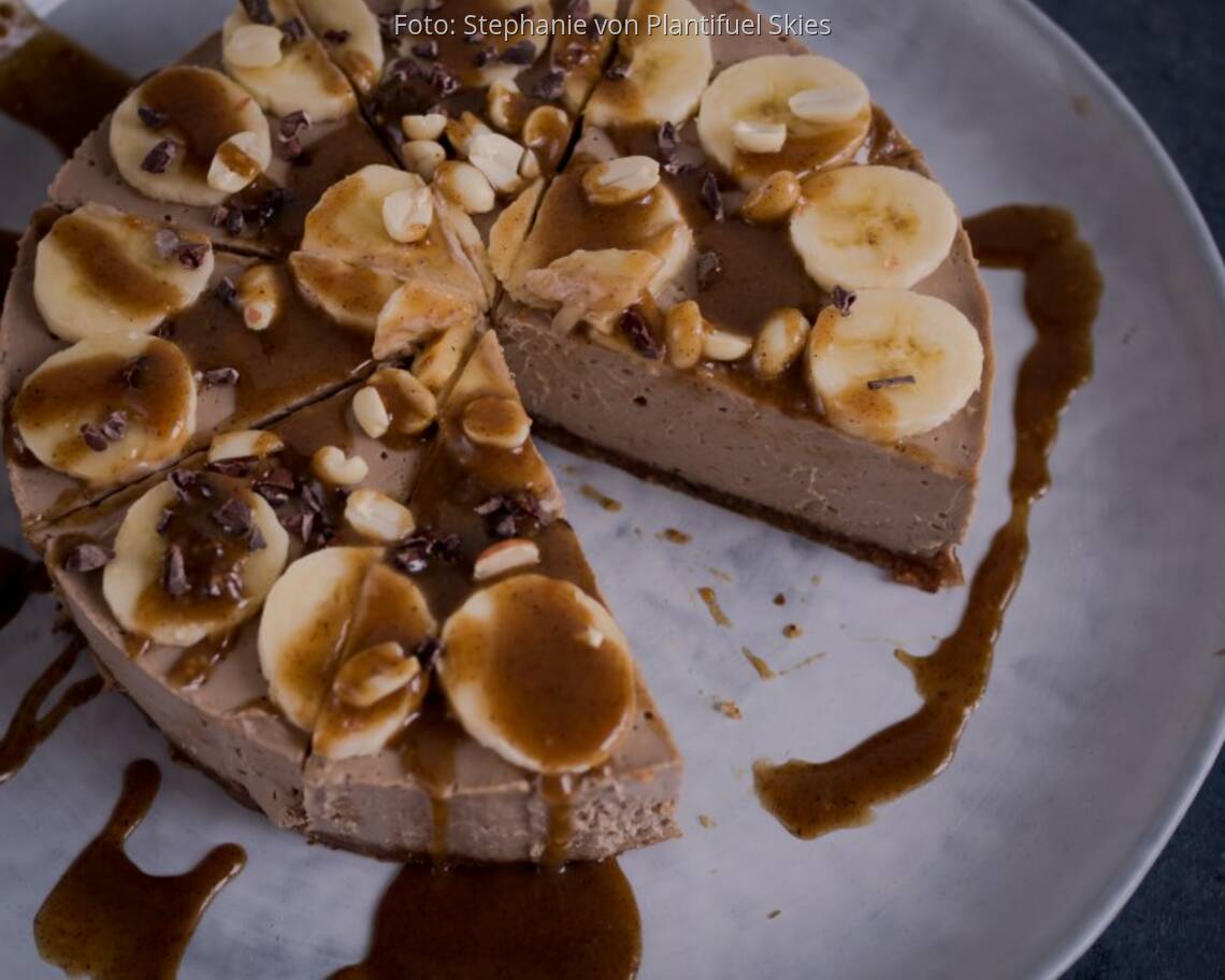 Torte aus Erdnussbutter auf großer heller Platte, dekoriert mit Bananenscheiben, Pinienkernen und Karamell