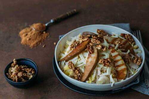 Fenchel-Chicoree-Salat mit karamellisierten Birnen neben Zimt und Walnüssen vor dunklem Hintergrund.