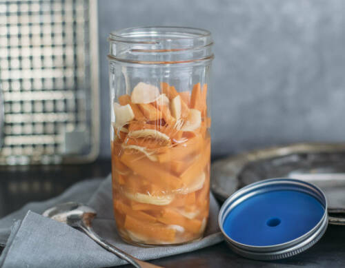 Fermentierte Ingwer-Karotten in offenem Weckglas neben Esslöffel vor hellem Hintergrund.