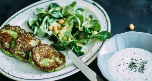 Brokkoli-Parmesan-Bratlinge auf einem Teller, dazu Feldsalat. Daneben steht ein Joghurtdip. Von oben fotografiert vor dunkelgrauem Hintergrund.