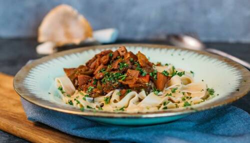 Pflanzliches Jackfruit-Gulasch mit Bandnudeln in einem hellblauen Teller mit goldenem Rand. Darüber Petersilie, im Hintergrund ein Löffel, von der Seite fotografiert.