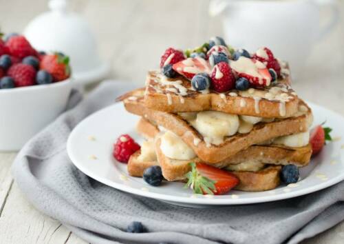 Stapel French Toast mit Bananen, Vanille Sauce und Beeren garniert vor hellem Hintergrund, von vorne fotografiert.