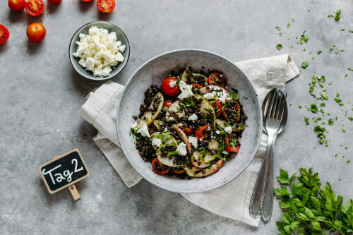 Fruchtiger Linsensalat in Schüssel neben Tomaten, Kräutern und Fetakäse vor hellem Hintergrund.