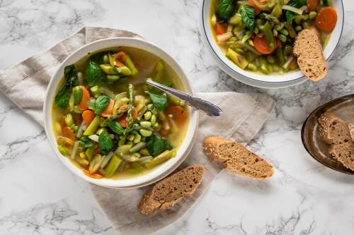 Frühlingsminestrone in Schüssel neben Dinkelbaguette vor hellem Hintergrund.