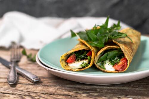 Zwei gerollte Pfannkuchen auf einem Teller, gefüllt mit Löwenzahn, Tomaten und Mozarella. Daneben Besteck, von der Seite fotografiert.