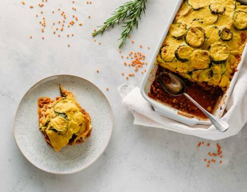 Gemüse-Linsen-Auflauf in Auflaufform und Teller neben Rosmarinzweig vor hellem Hintergrund.