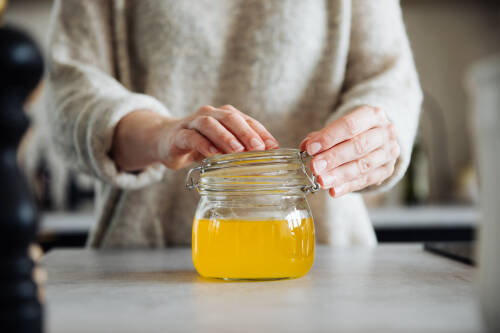 Ghee im Schraubglas und Händen vor hellem Hintergrund.
