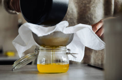 Ghee in Weckglas mit Topf und Sieb in Händen vor hellem Hintergrund.