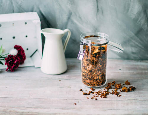 Granola-Knusper-Müsli in Glas neben Milchkanne vor hellem Hintergrund.
