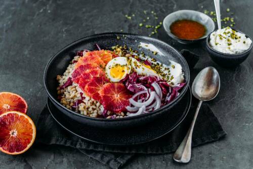 Graupen-Powerbowl in schwarzer Schüssel neben Blutorangen und Gewürzen vor dunklem Hintergrund.