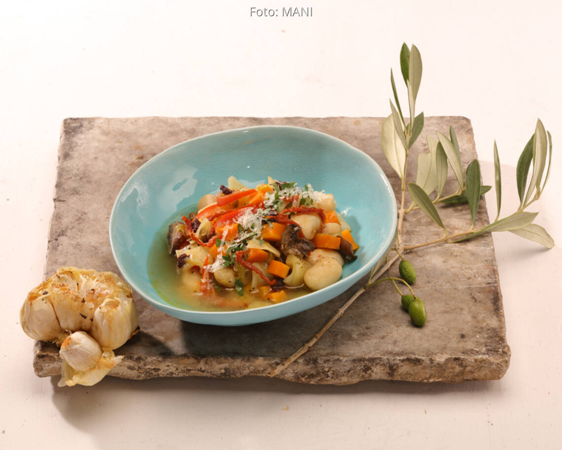 Bohnen-Eintopf in blauer Schale auf Holzbrett mit gebackenem Knoblauch und Olivenzweig