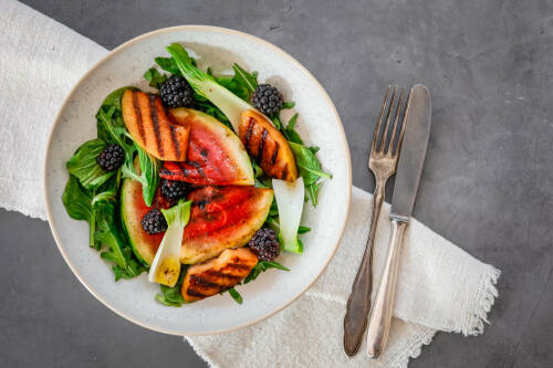 Grillmelone mit Salat auf Teller neben Besteck vor hellem Hintergrund.